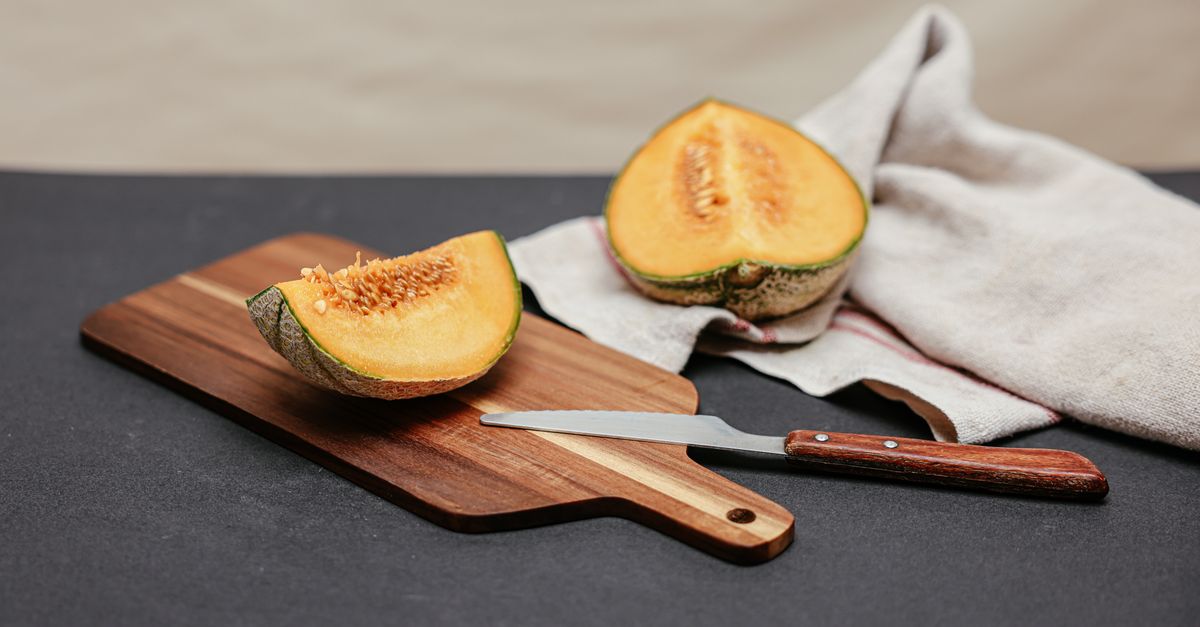 Un quartier de melon jaune coupé et une moitié de melon posés sur une planche à découper en bois, avec un couteau et un torchon en lin à proximité.