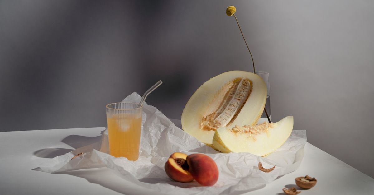 Un demi-melon coupé et une tranche posés sur du papier blanc à côté d'un verre de jus avec paille et d'une pêche coupée en deux.