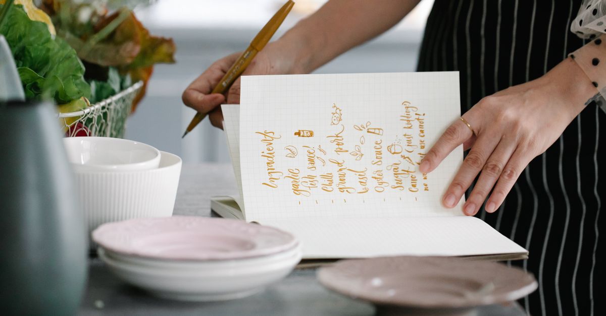 Une femme en tablier rayé écrit une liste d'ingrédients à la main dans un carnet, entourée de bols et assiettes en céramique sur une table de cuisine.