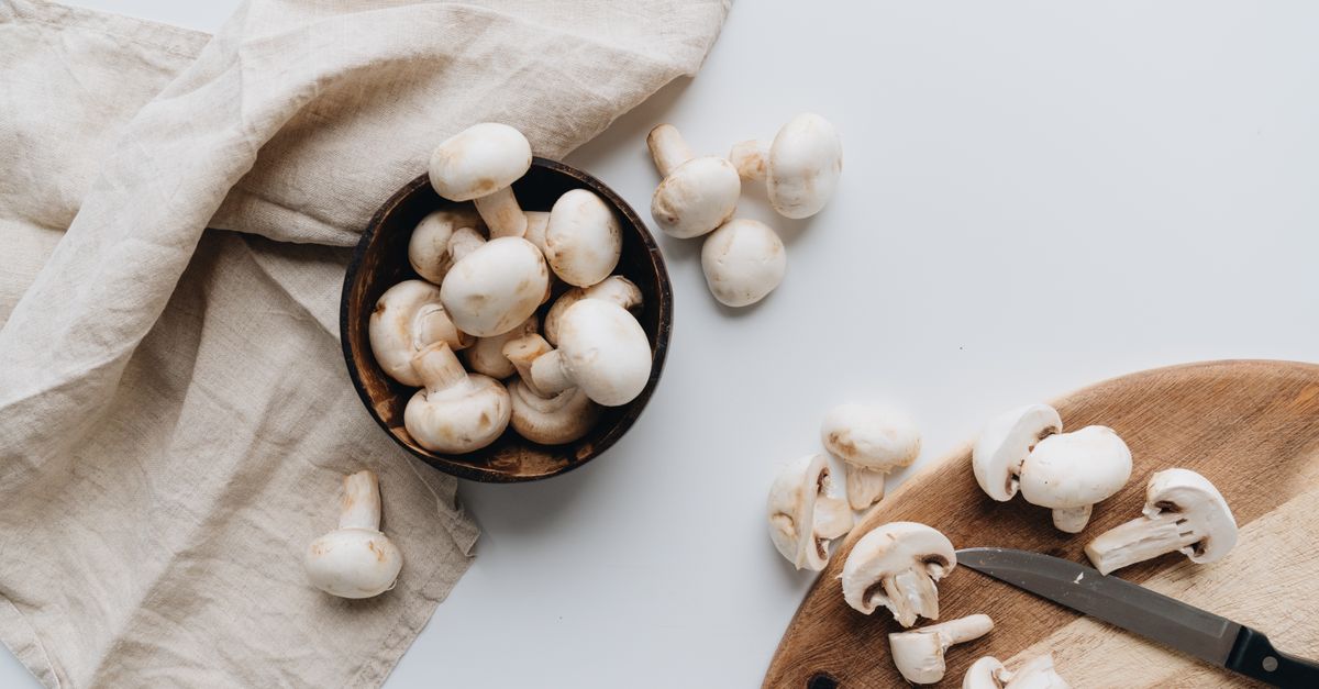 Champignons de Paris frais entiers et coupés en tranches sur une planche en bois, avec un couteau et un torchon en lin à côté.