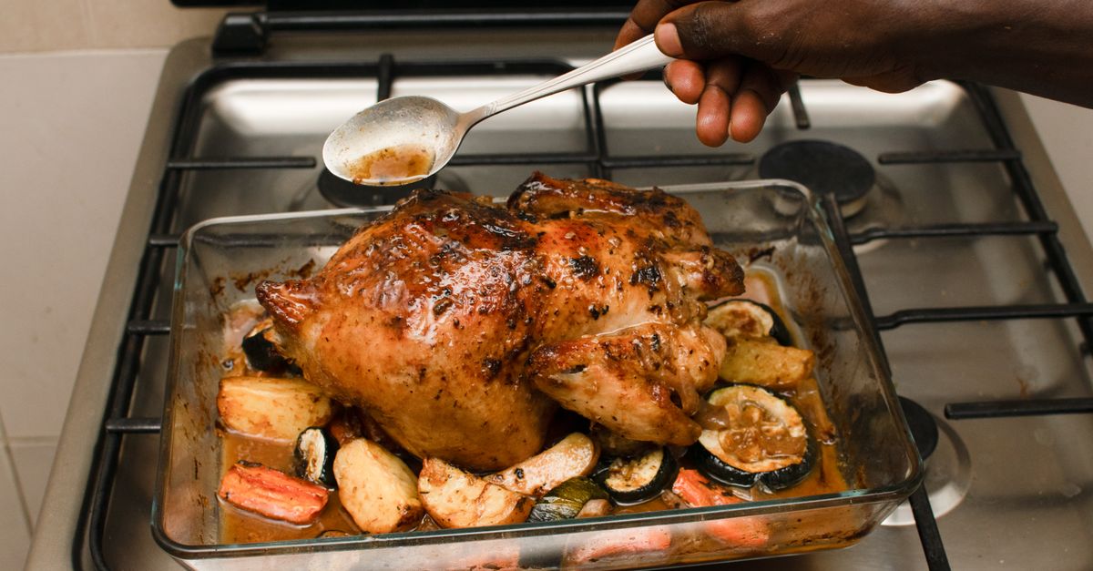 Une main arrose au moyen d'une cuillère un poulet entier rôti disposé dans un plat avec des légumes, posé sur une cuisinière à gaz.