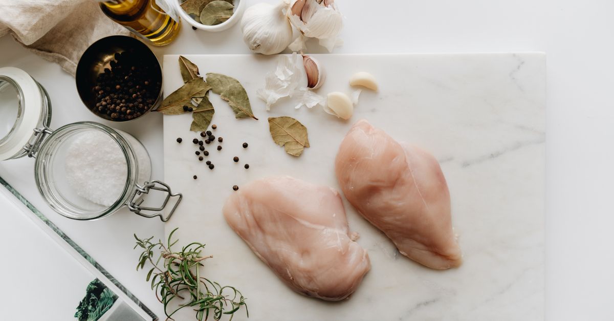 Deux blancs de poulet crus posés sur une planche en marbre blanc, entourés d'ail, de romarin, de feuilles de laurier et d'épices.
