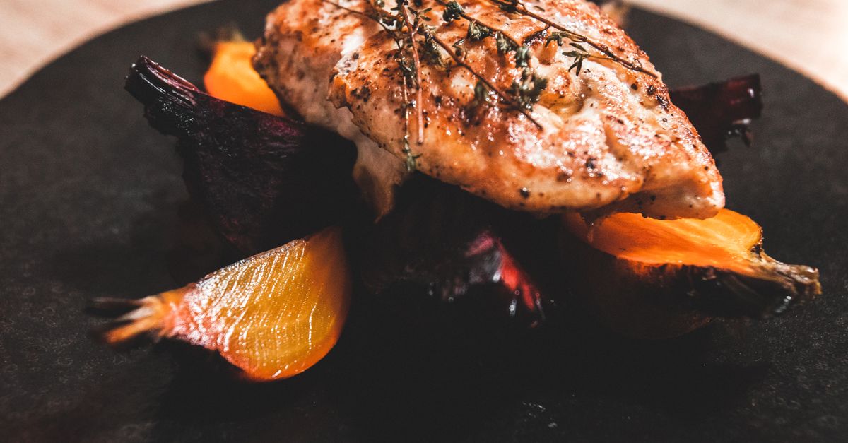 Un filet de poulet doré garni de thym, servi sur un lit de légumes racines rôtis dans une assiette noire.