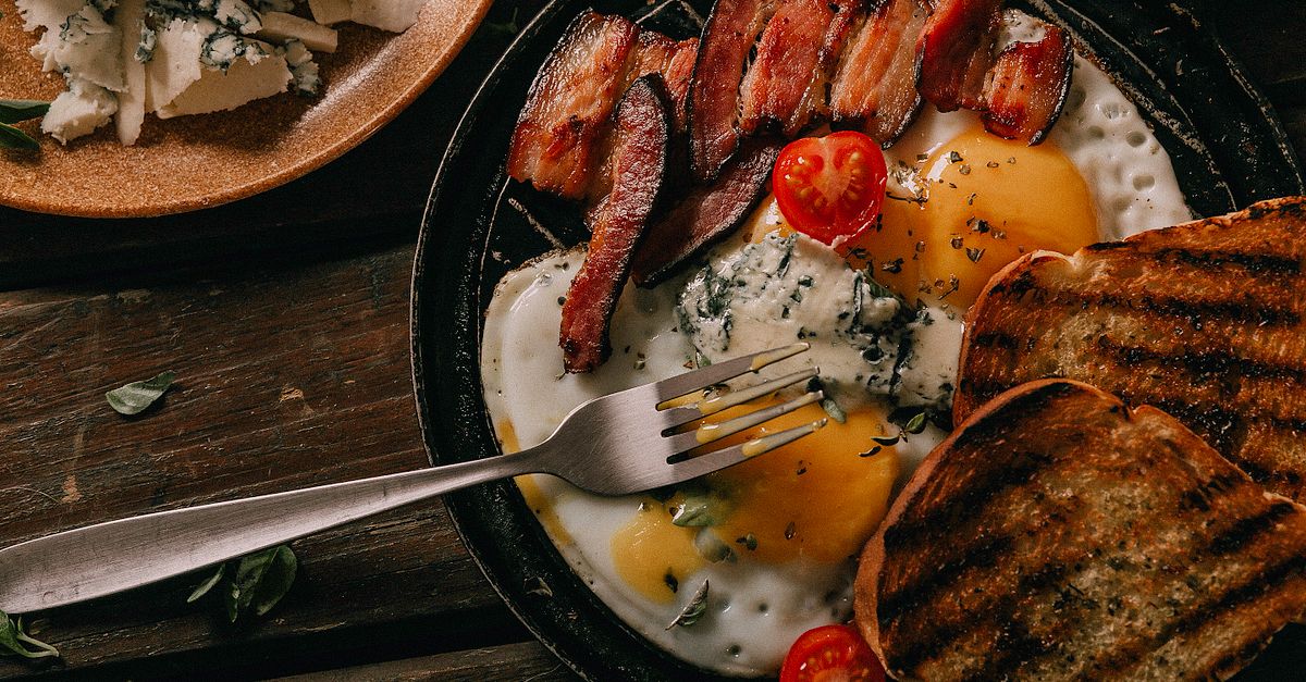 Œufs au plat cuits dans une poêle en fonte, servis avec bacon grillé, tomate cerise, fromage bleu et pain grillé, jaune bien coulant.