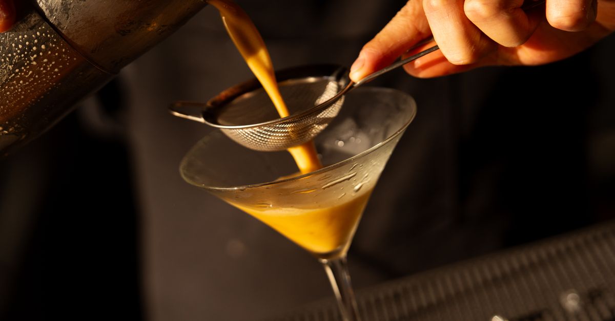 Une passoire fine en métal filtre un liquide doré versé dans un verre à cocktail.