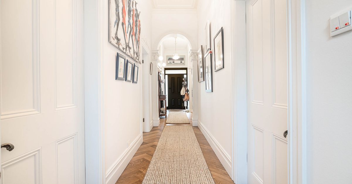 Un couloir étroit et lumineux aux murs blancs, décoré de cadres et éclairé par un lustre, avec un tapis de couloir au sol.