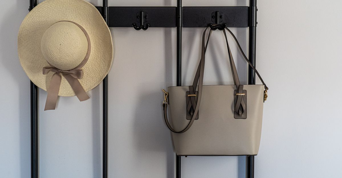 Un chapeau de paille et un sac à main en cuir beige suspendus à une patère murale noire fixée sur un mur blanc.