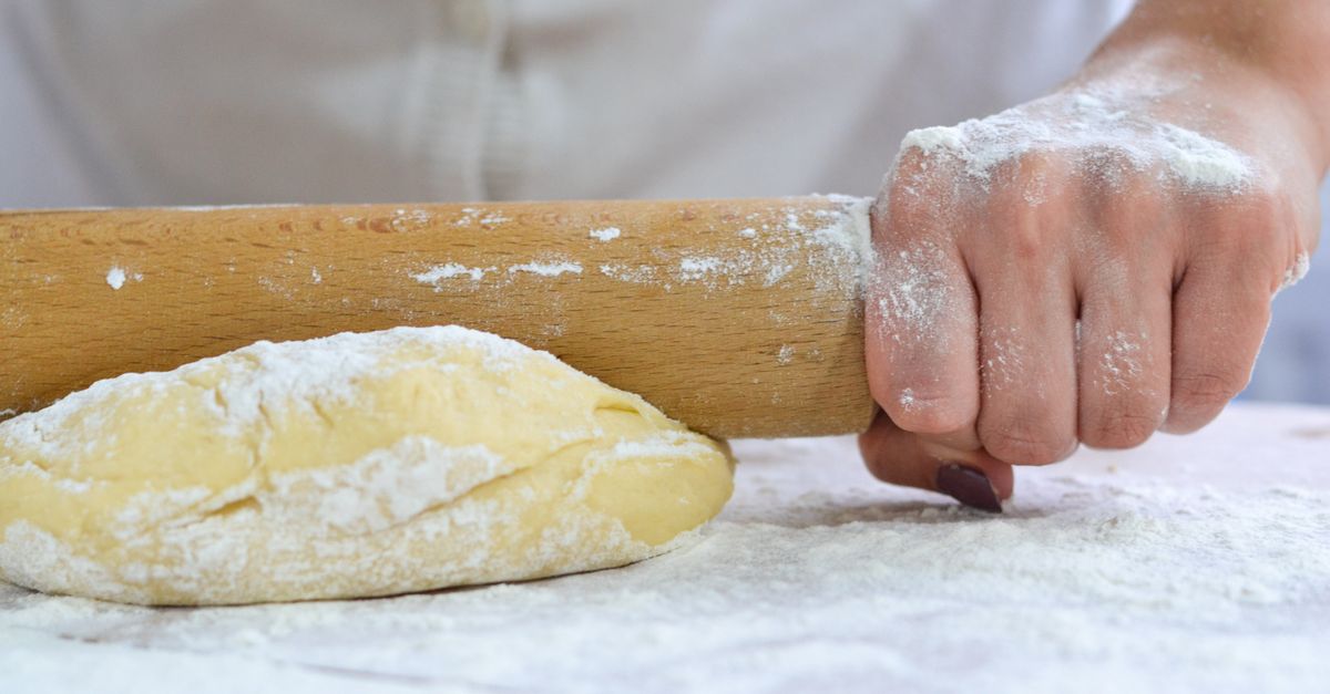 Une main farinée tient un rouleau à pâtisserie en bois posé sur un pâton de pâte brisée farinée, prêt à être étalé.