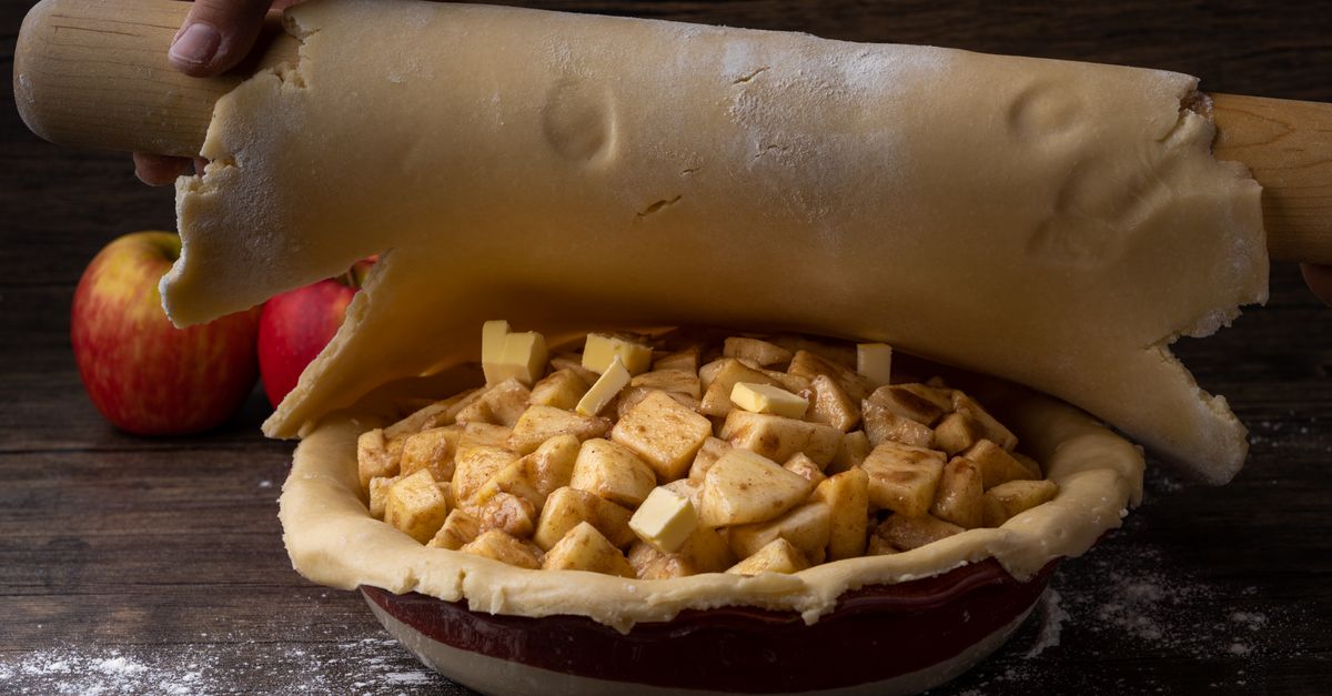 Une pâte étalée sur un rouleau à pâtisserie en bois est déposée sur un plat rempli de pommes coupées, prête à recouvrir une tarte.