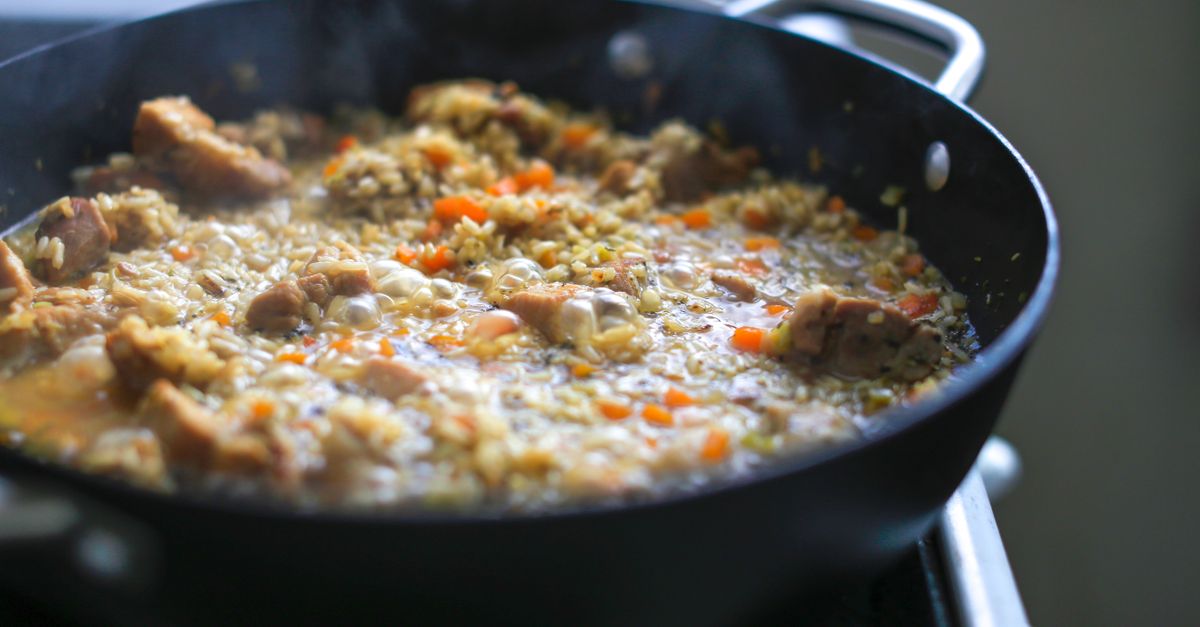 Une poêle noire sur le feu contenant de la viande en morceaux mijotant dans un liquide de cuisson avec des carottes et du riz.