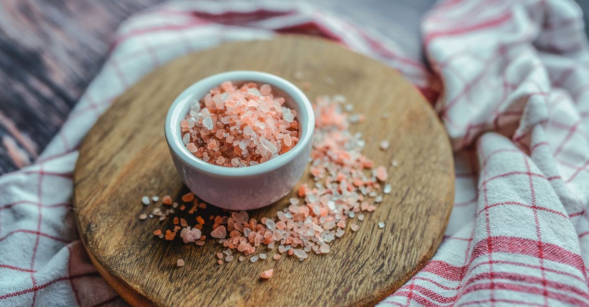 Un bol de sel rose de l'Himalaya posé sur une planche à découper en bois ronde, avec des grains de sel épars autour et un torchon à carreaux rouge et blanc en arrière-plan.