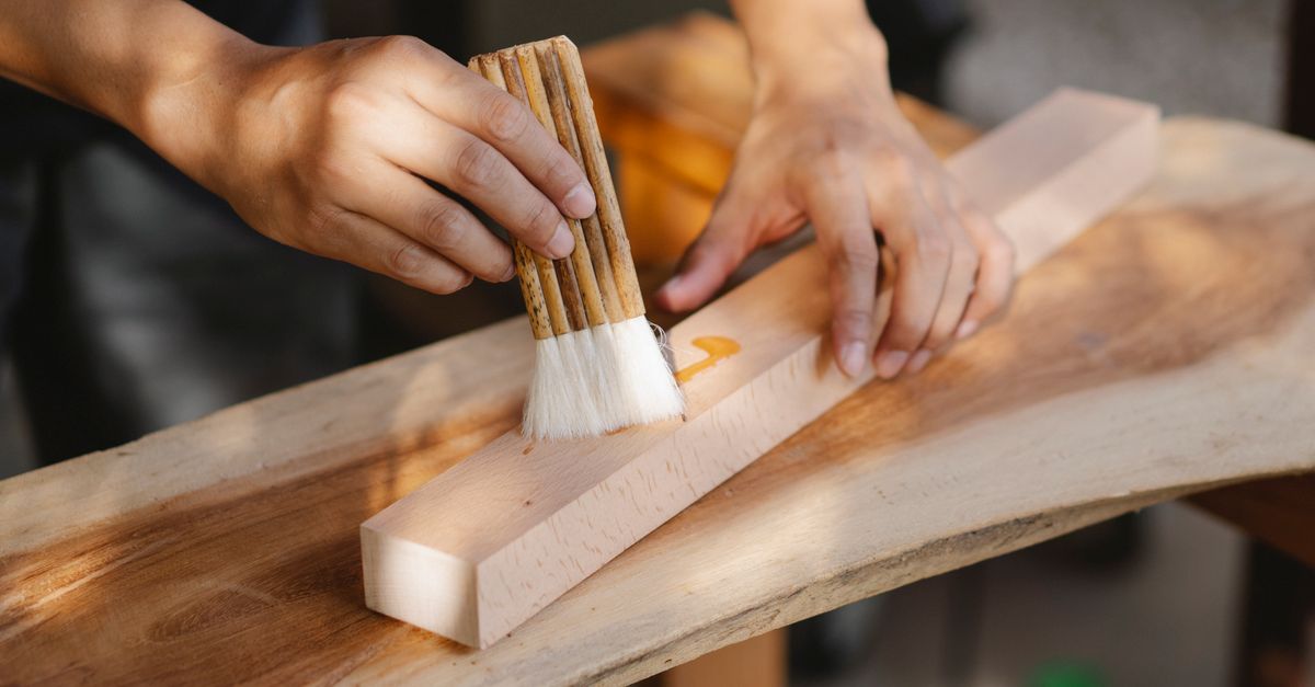 Deux mains appliquent de l'huile au pinceau sur une pièce de bois brut posée sur une planche.