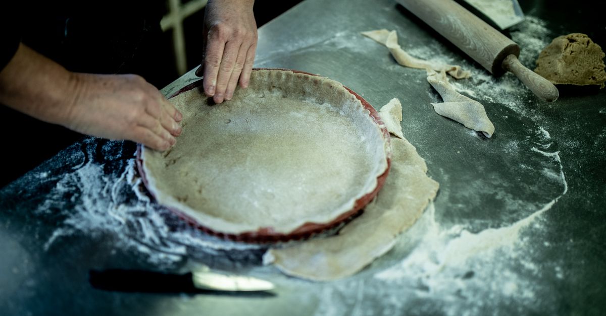 Des mains façonnent une pâte brisée crue dans un moule à tarte posé sur un plan de travail fariné, avec un rouleau à pâtisserie à proximité.