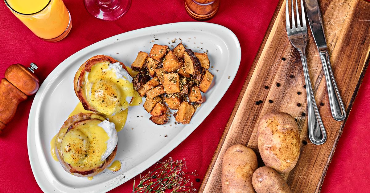 Œufs bénédicte nappés de sauce hollandaise dorée sur une assiette blanche, accompagnés de pommes de terre rôties.