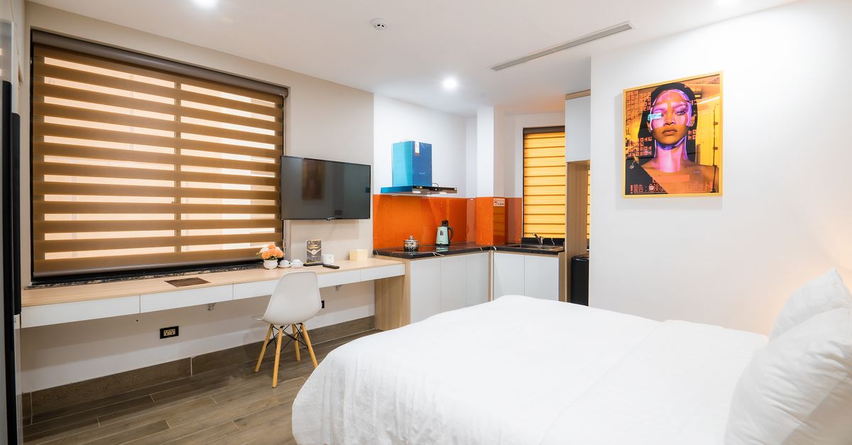 Studio lumineux avec lit blanc au premier plan, bureau, kitchenette et grande fenêtre à volets bois en fond.