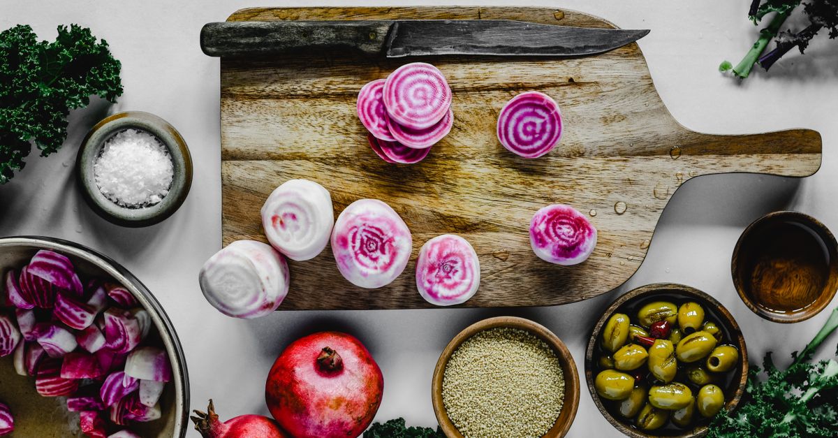 Des rondelles de betteraves chioggia roses et blanches tranchées sur une planche à découper en bois, entourées de bols d'olives, de quinoa et de grenade.