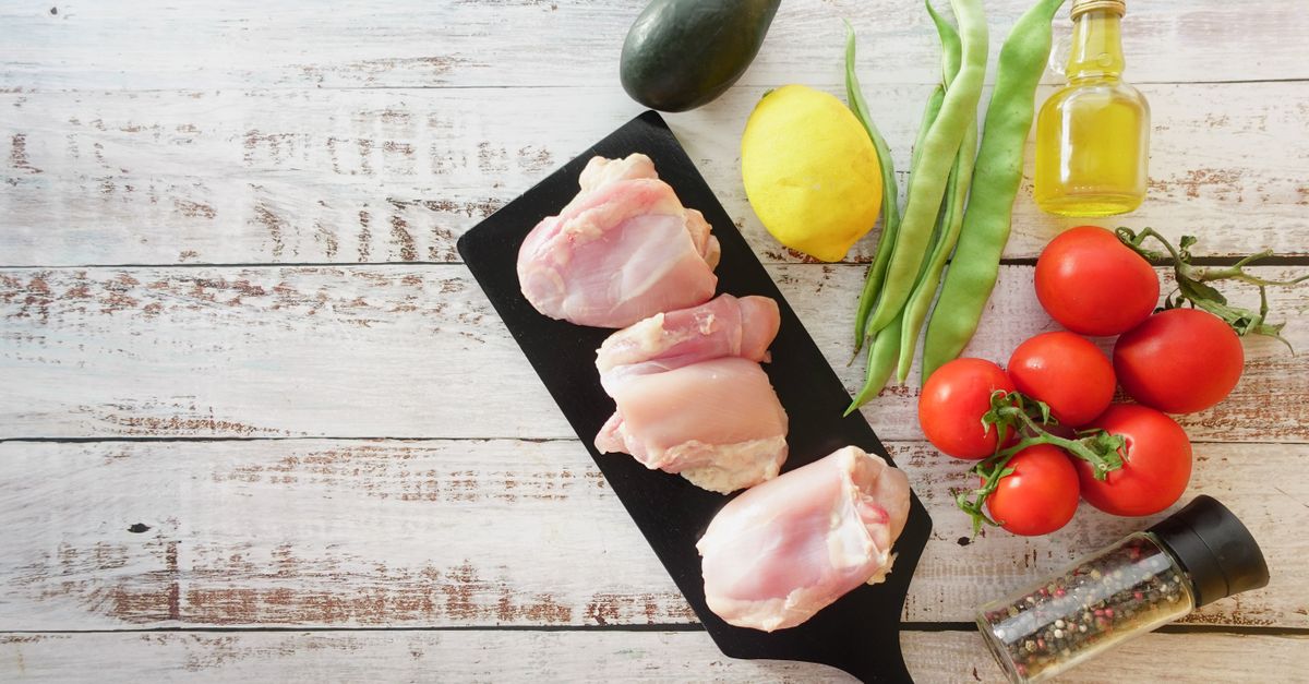 Des morceaux de poulet cru disposés sur une planche noire, entourés de citron, tomates, haricots verts et huile d'olive.