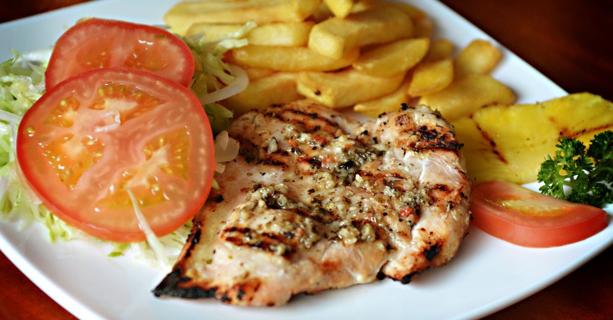 Un blanc de poulet grillé marqué au barbecue, servi sur une assiette blanche avec des frites, de la salade et des tranches de tomate.