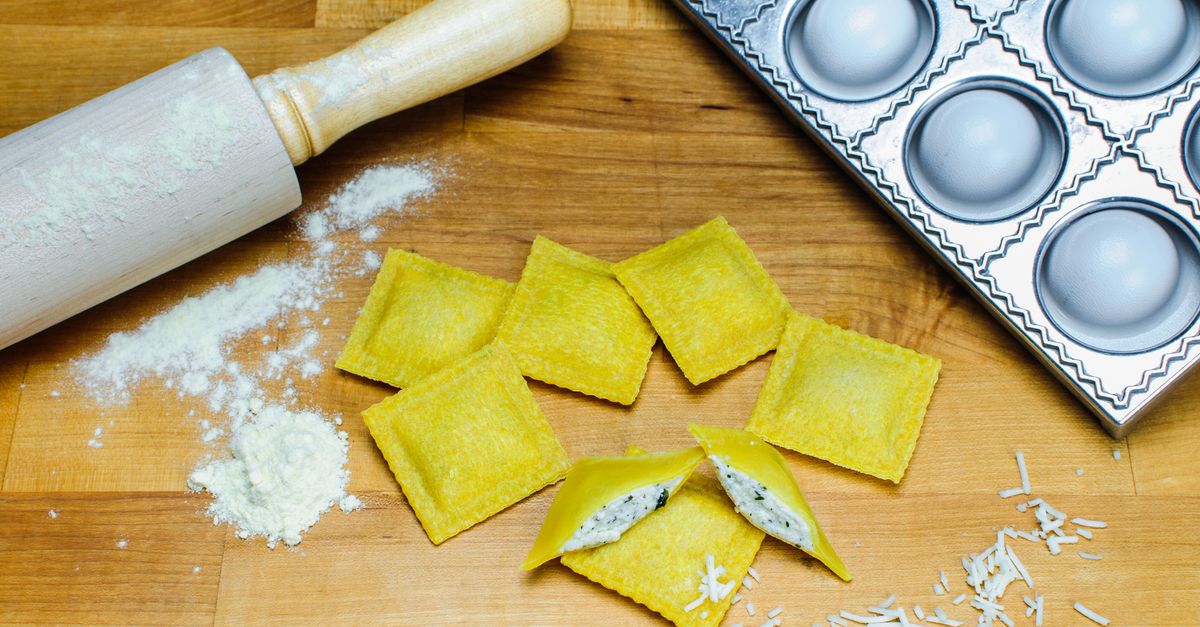 Des raviolis jaunes farcis disposés sur un plan de travail en bois, entourés de farine, d'un rouleau à pâtisserie et d'un moule à raviolis, l'un d'eux ouvert révélant la farce.