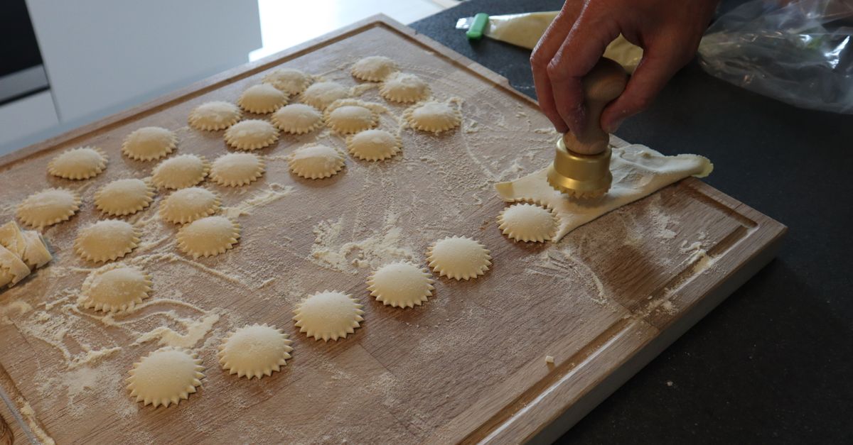 Une main utilise un emporte-pièce pour découper des raviolis fourrés disposés en rangées sur une planche en bois farinée.