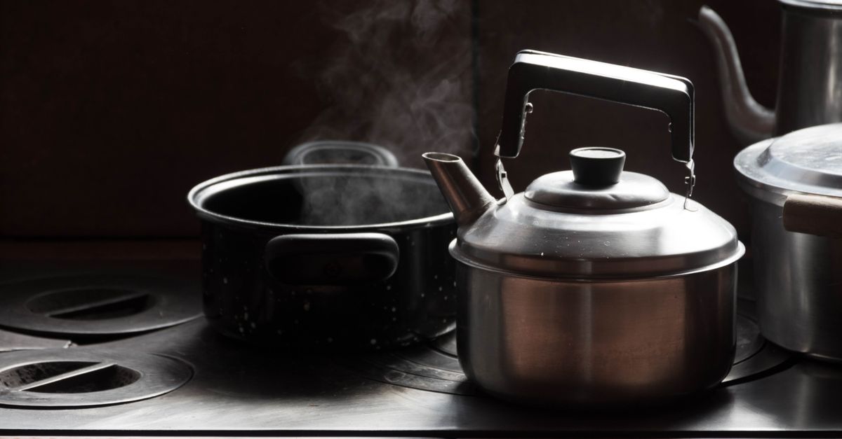 Une casserole en métal noir fume sur une cuisinière à côté d'une bouilloire en inox, évoquant un liquide en train de mijoter.