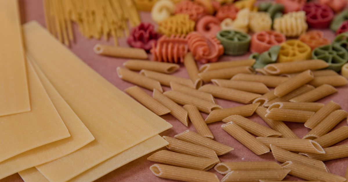 Assortiment de pâtes sèches crues de formes et couleurs variées disposées sur une surface rose.
