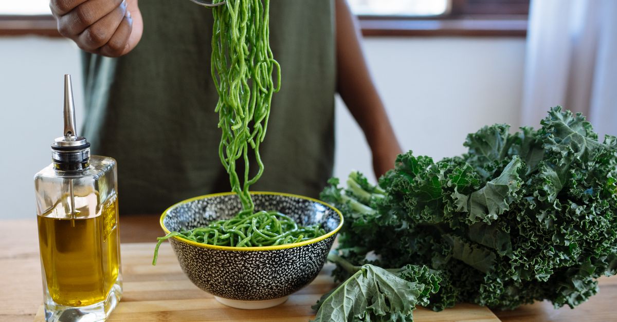 Une personne soulève des pâtes vertes cuites au-dessus d'un bol en céramique, avec une bouteille d'huile d'olive et du chou kale à côté sur une table en bois.