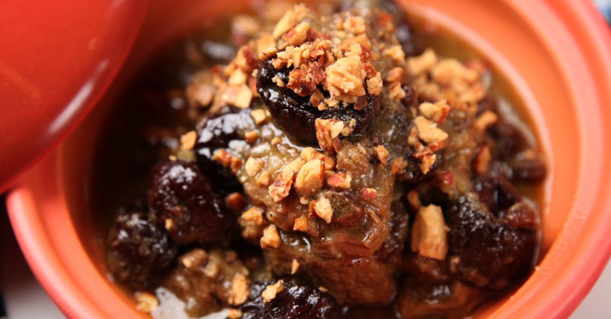 Un tajine en terre cuite orange contenant de la viande mijotée nappée de sauce et parsemée d'amandes concassées et de pruneaux.