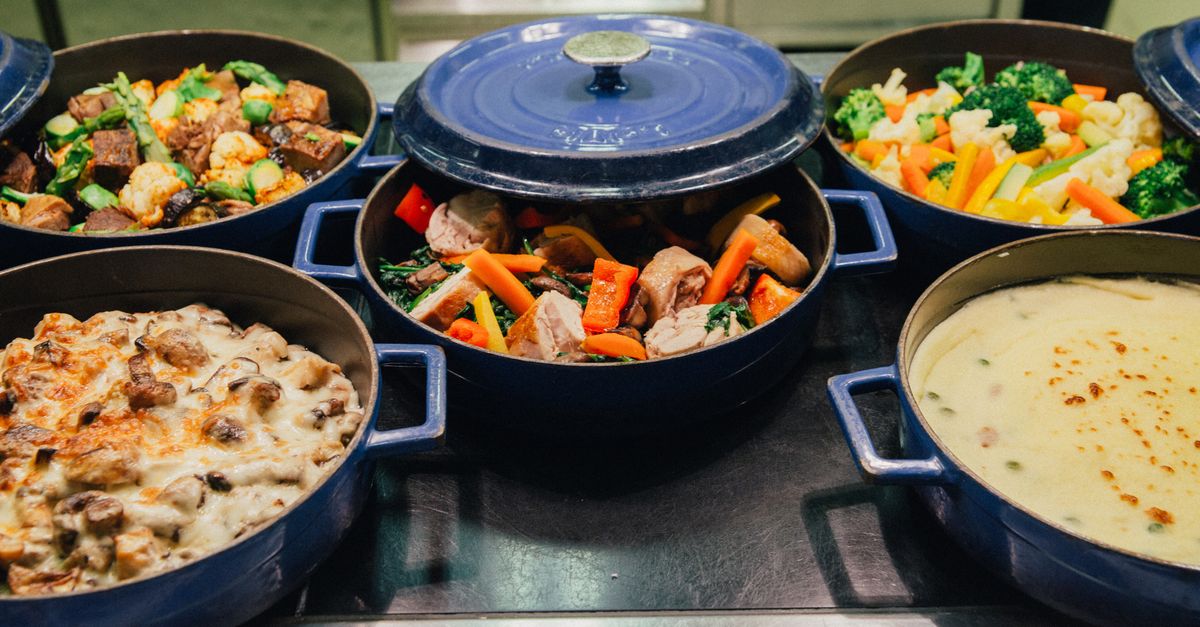 Une cocotte en fonte bleue ouverte laisse voir un mijoté de viande et de légumes (carottes, poireaux) entouré d'autres plats sur un comptoir.