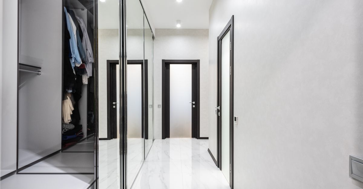 Une armoire avec porte miroir pleine hauteur reflète un couloir aux portes noires dans une pièce claire.
