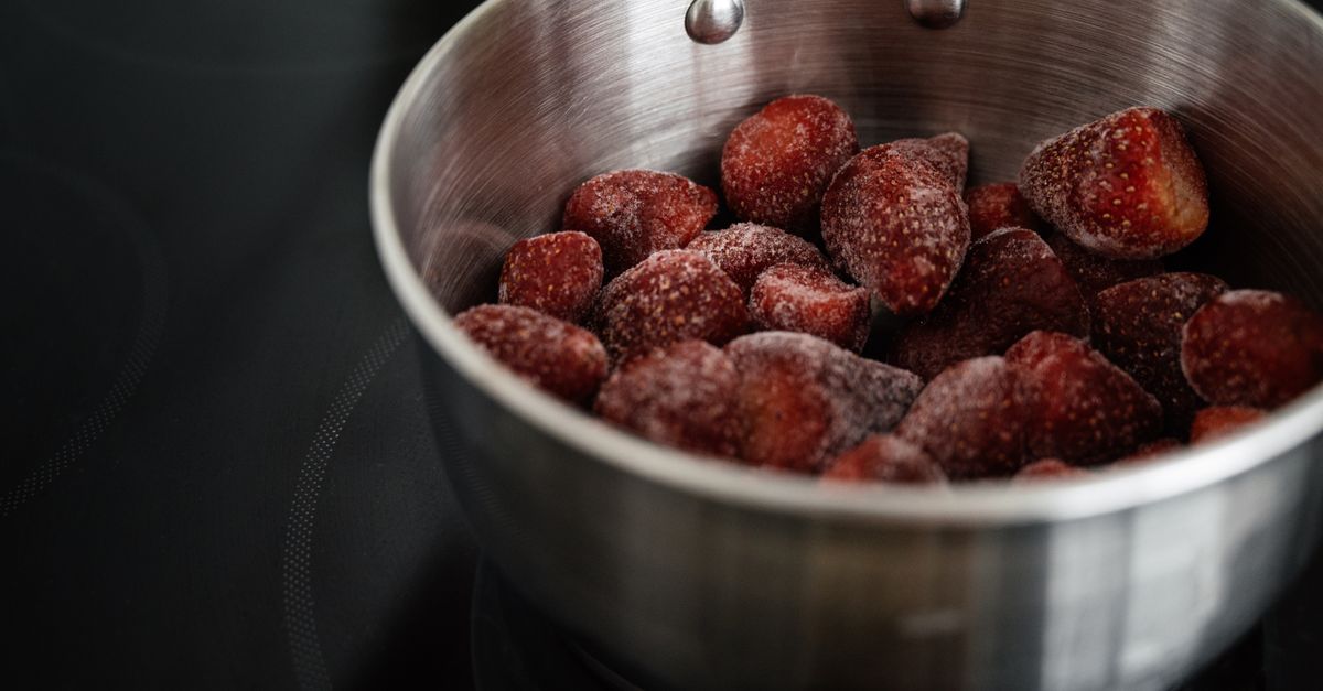 Des fraises congelées, couvertes de givre, entassées dans une casserole en inox posée sur une plaque de cuisson noire.