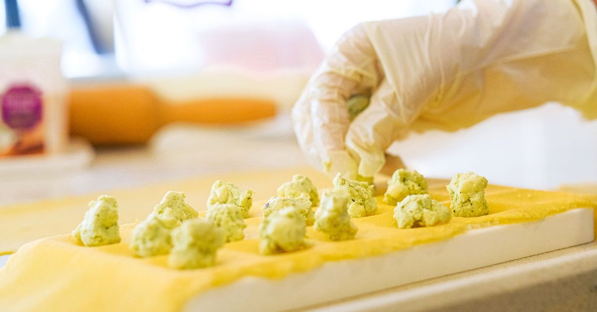Une main gantée dépose des tas de farce verte sur une pâte fraîche jaune avant de former des raviolis faits maison.