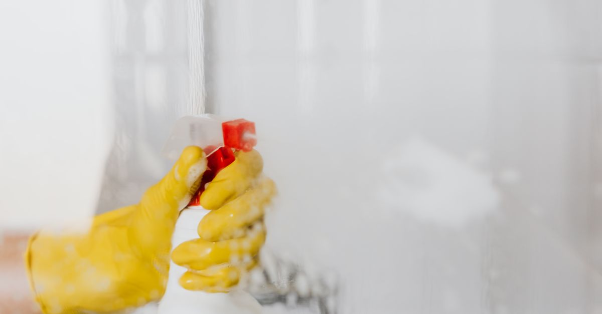 Une personne portant des gants en caoutchouc jaune nettoie du carrelage avec un spray et de la mousse dans une salle de bain.