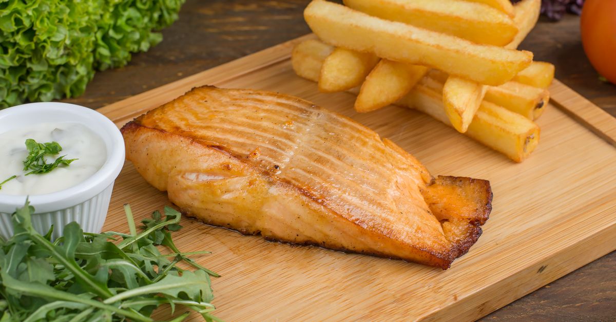 Un filet de saumon cuit avec la peau bien dorée, servi sur une planche en bois avec frites, roquette et sauce blanche.