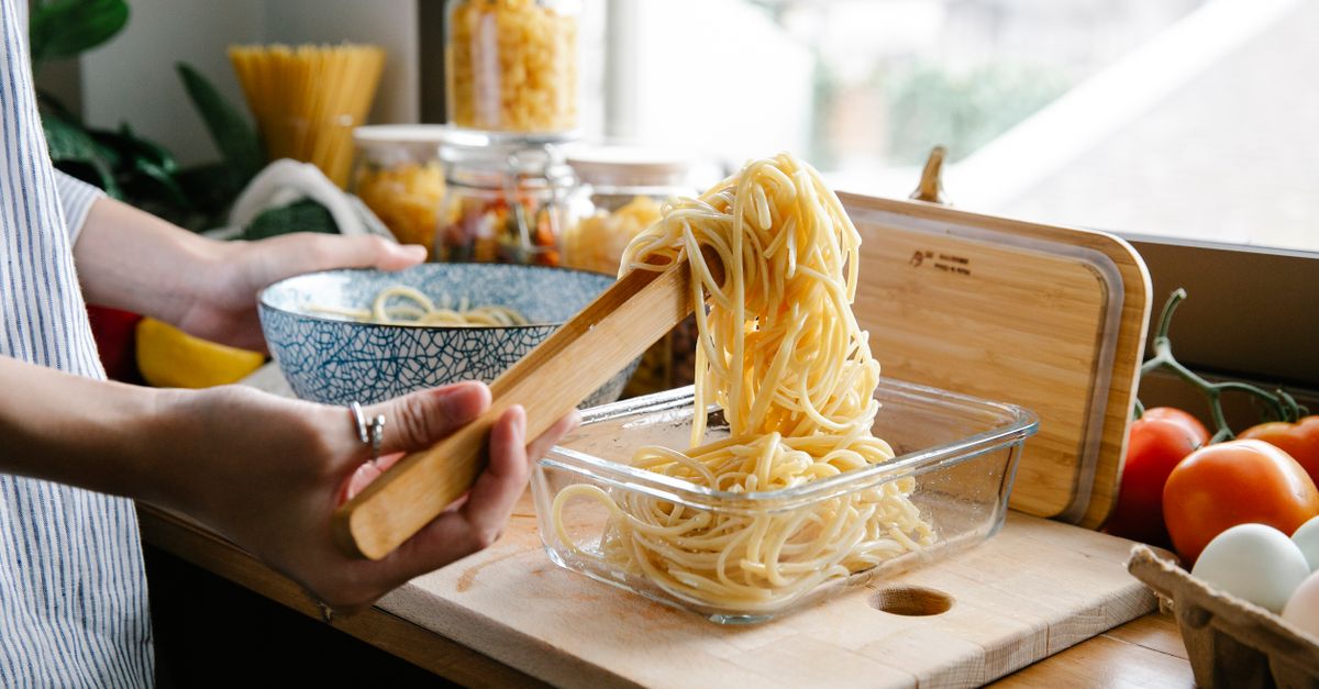 Une personne transfère des spaghettis cuits à l'aide de pinces en bois dans une boîte en verre pour la conservation, sur un plan de travail en bois.