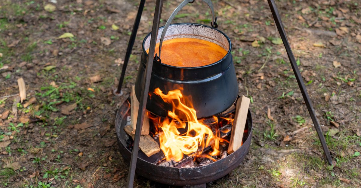 Une marmite en fonte noire suspendue au-dessus d'un feu de bois contient une sauce mijotée orangée qui bouillonne doucement.