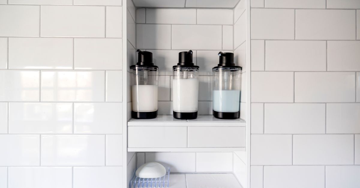 Niche de douche carrelée en blanc avec trois distributeurs de savon liquide et un porte-savon vide en bas.