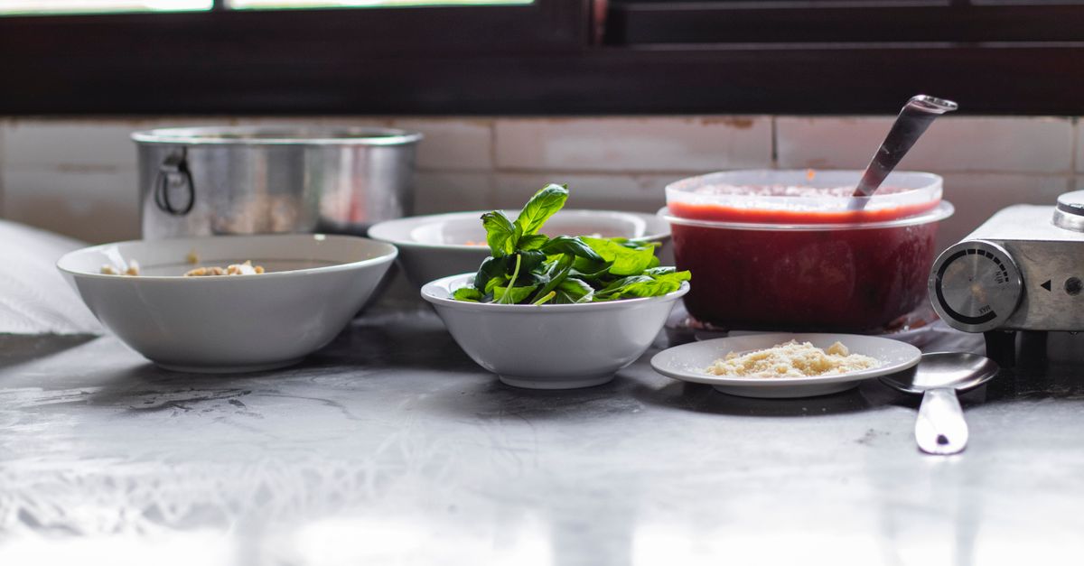 Un bol de sauce tomate maison posé sur un plan de travail en marbre, entouré de bols de basilic frais et de parmesan râpé.