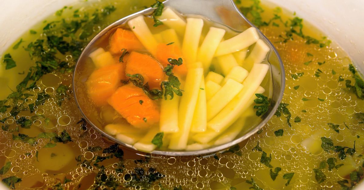 Une louche servant un bouillon clair avec des légumes coupés en morceaux et julienne dans une assiette creuse.