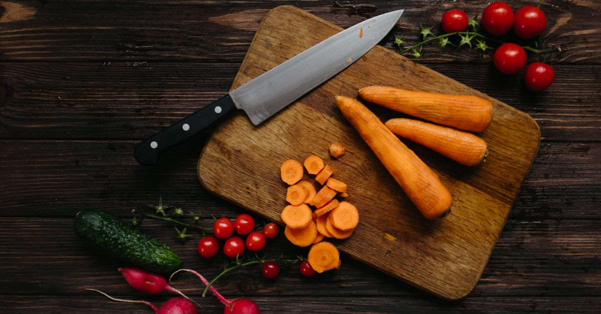 Des carottes entières et des rondelles de carotte fraîchement coupées sur une planche à découper en bois, avec un couteau, des tomates cerises et un radis autour.