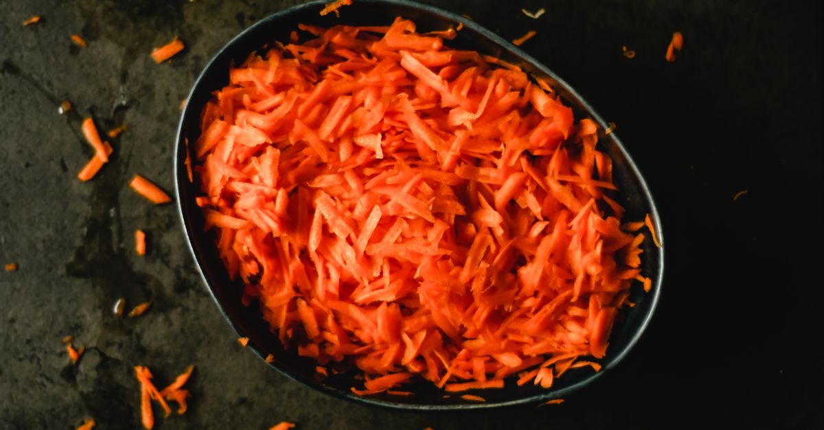 Des carottes râpées orange vif sont présentées dans un bol foncé, avec quelques brins éparpillés sur la surface sombre autour.