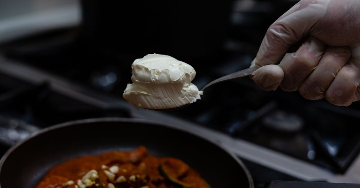 Une cuillère de crème épaisse tenue au-dessus d'une poêle de légumes en sauce.