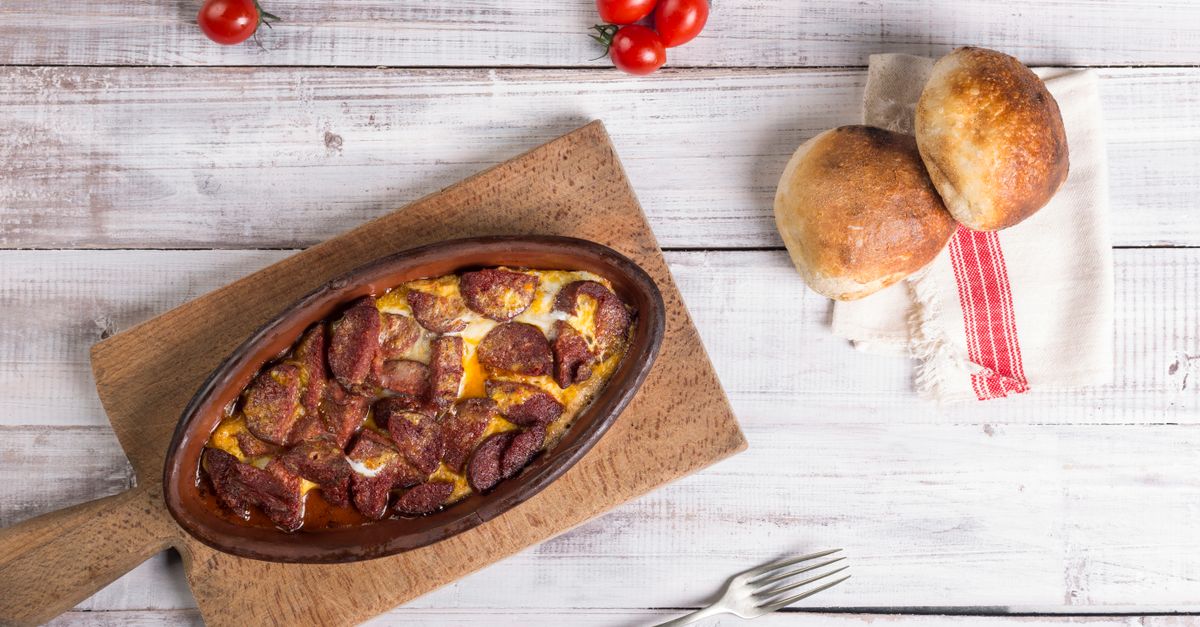 Un plat en terre cuite ovale avec des œufs cuits recouverts de morceaux de chorizo, posé sur une planche en bois à côté de petits pains et de tomates cerises.