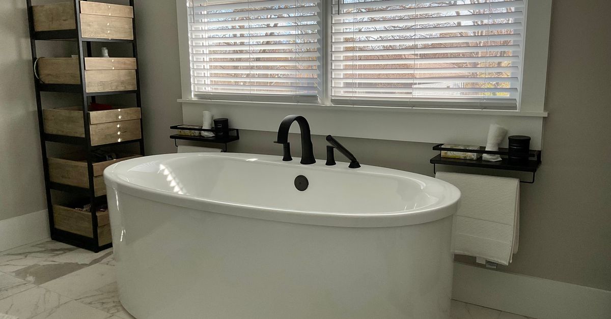 Baignoire blanche autonome avec deux petites étagères noires fixées au mur de part et d'autre, surmontée d'une fenêtre à volets.