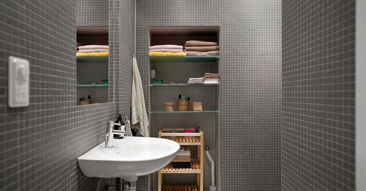 Étagères en verre encastrées au-dessus d'un lavabo, garnies de serviettes pliées et de produits de salle de bain, dans une salle de bain carrelée de mosaïque grise.