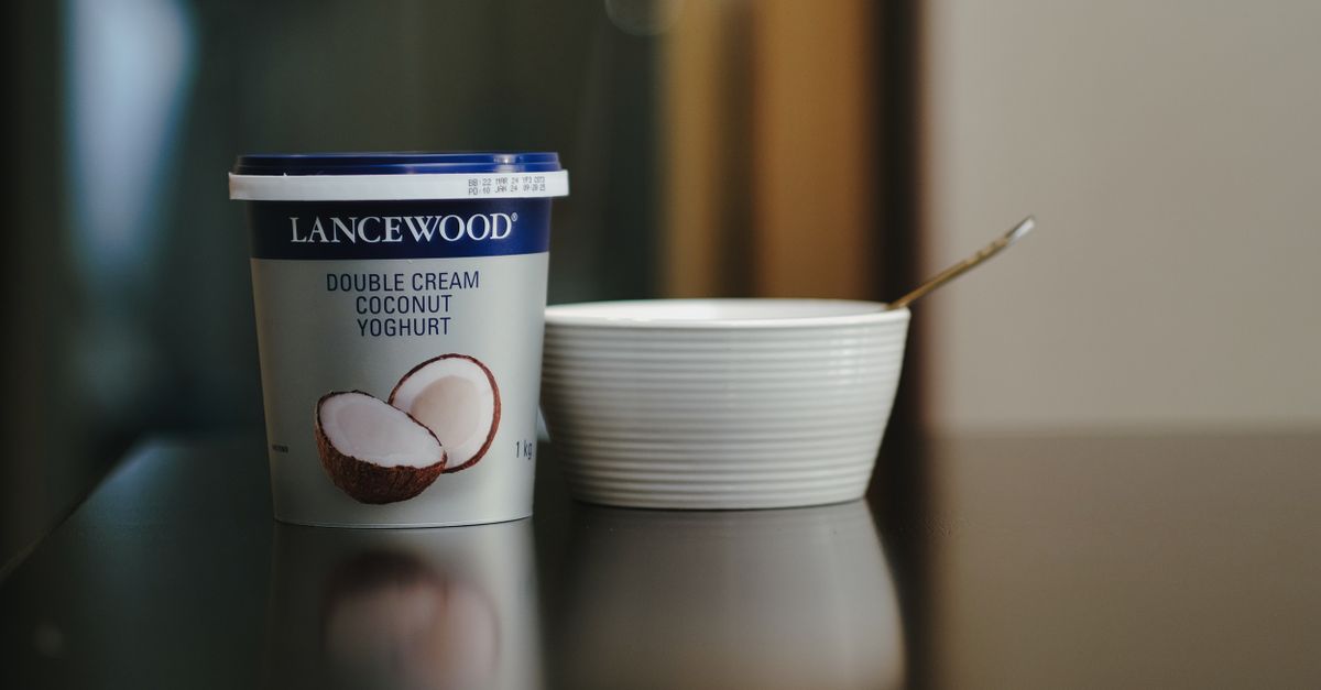 Un pot de yaourt à la noix de coco de marque Lancewood posé sur une table sombre à côté d'un bol en céramique blanche vide avec une cuillère.