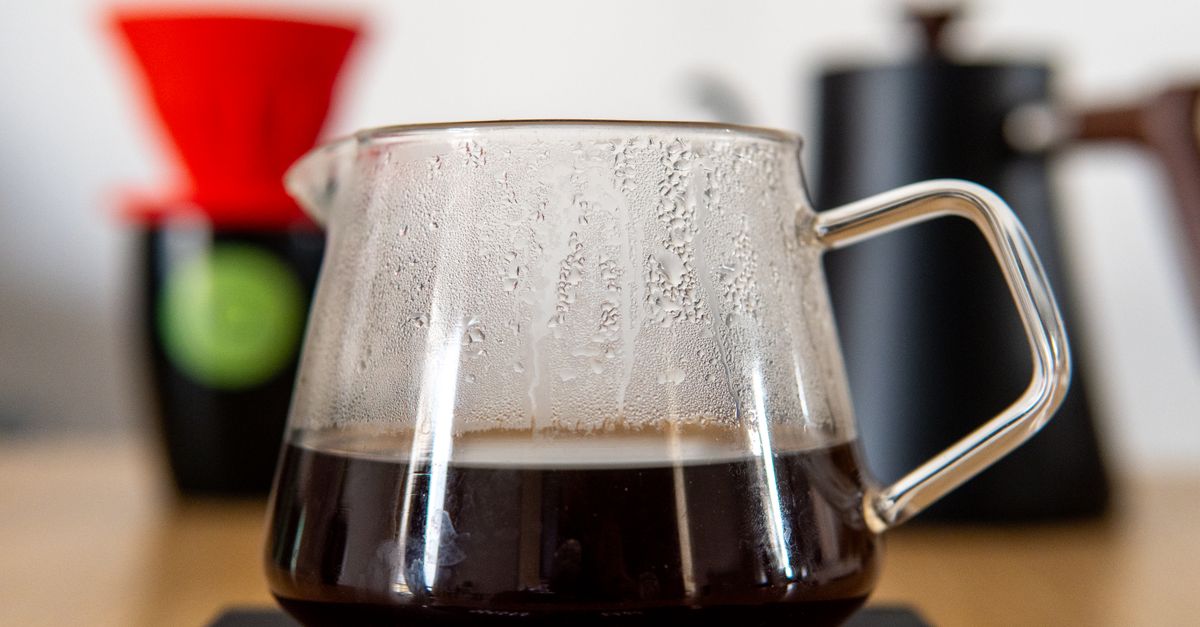 Une carafe en verre transparente remplie de café filtre posée sur une plaque chauffante noire, avec un porte-filtre rouge en arrière-plan.