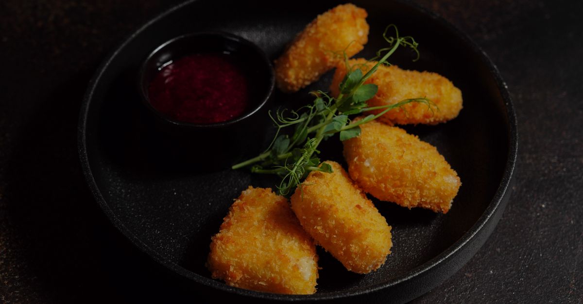 Cinq bâtons de mozzarella panés et frits, dorés et croustillants, servis sur une assiette noire avec une sauce rouge et quelques brins de verdure.