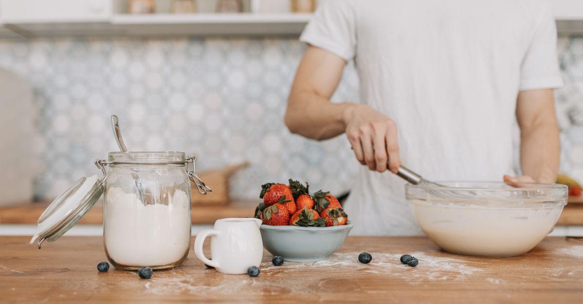 Une personne fouette une préparation lisse dans un bol en verre, à côté d'un pot de lait, de fraises et de myrtilles sur un plan de travail en bois.