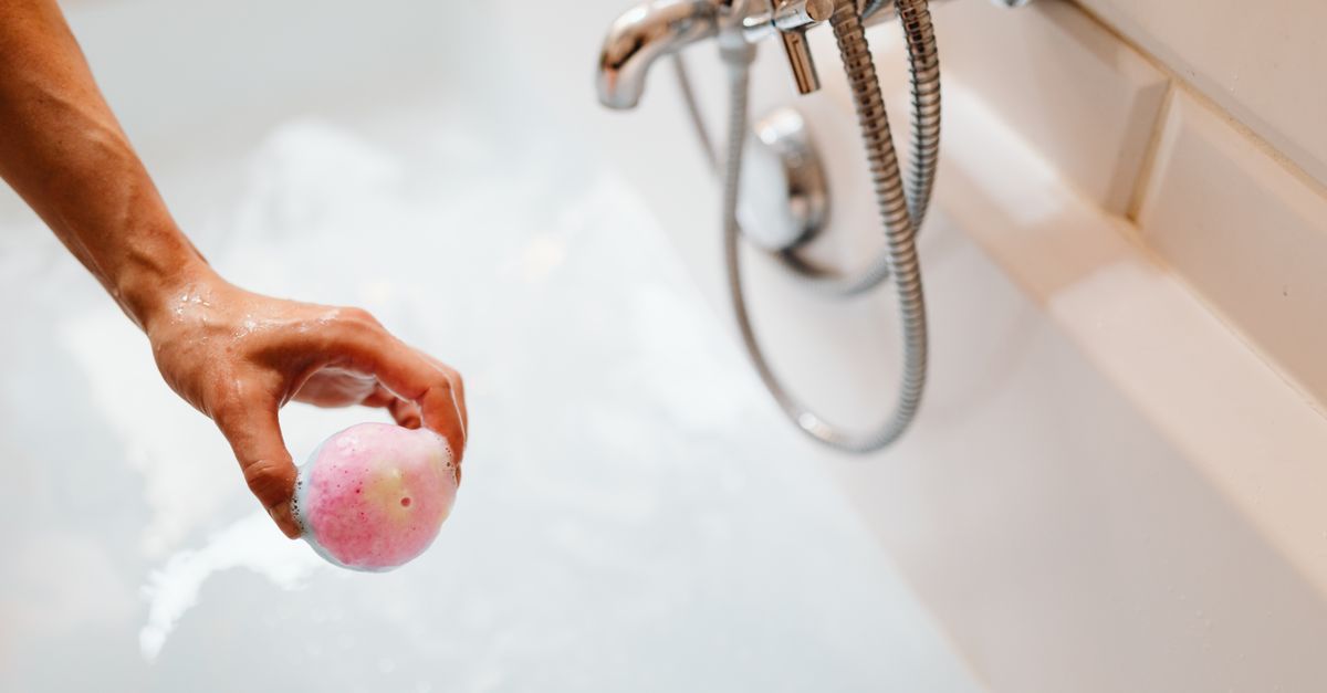 Une main tenant une boule de bain rose au-dessus d'une baignoire blanche remplie d'eau savonneuse, avec un robinet et une douchette en métal visibles.